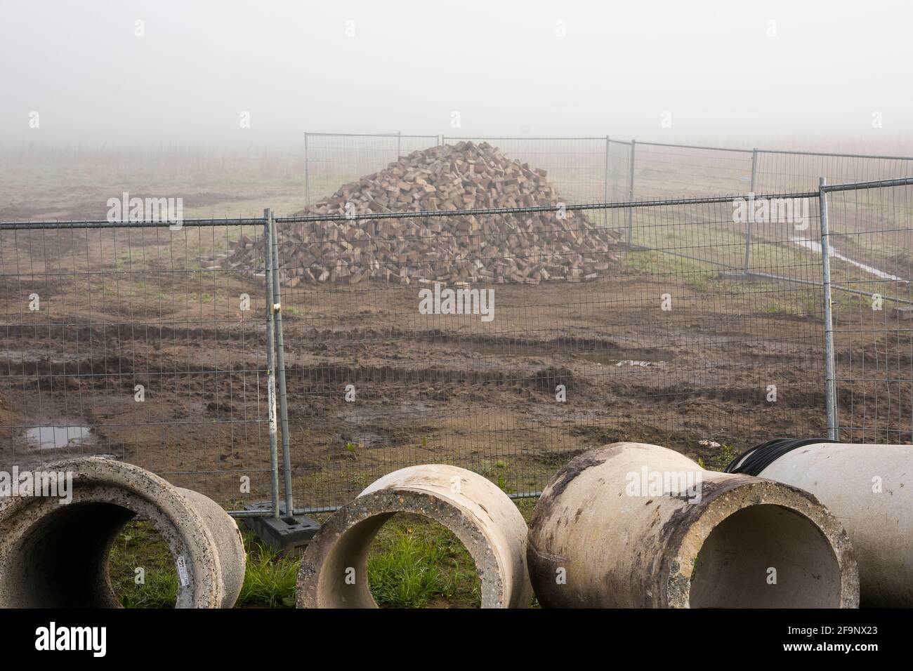 Sewer pipes and a stack of pavers behind a fence at building site in the mist, Netherlands Stock Photo