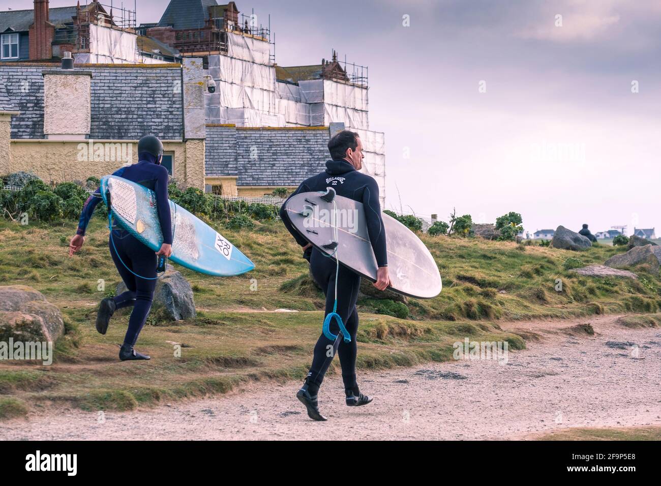 Two keen surfers carrying their surfboards and running along the coast path on the coast of Fistral in Newquay in Cornwall. Stock Photo