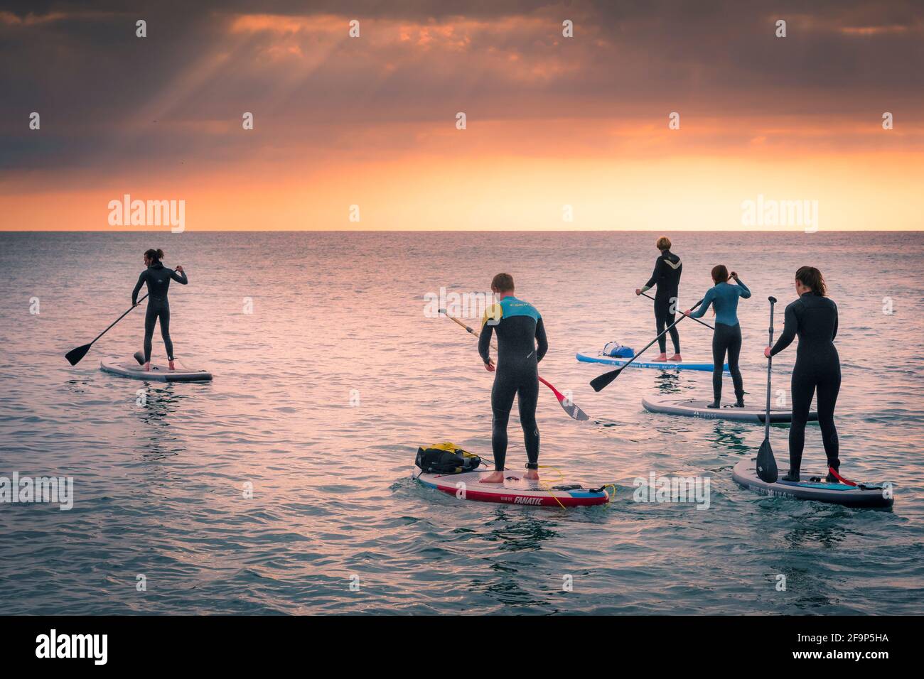 A group of holidaymakers on a staycation holiday paddling on Stand Up Paddleboards as the sun sets over Fistral Bay in Newquay in Cornwall. Stock Photo