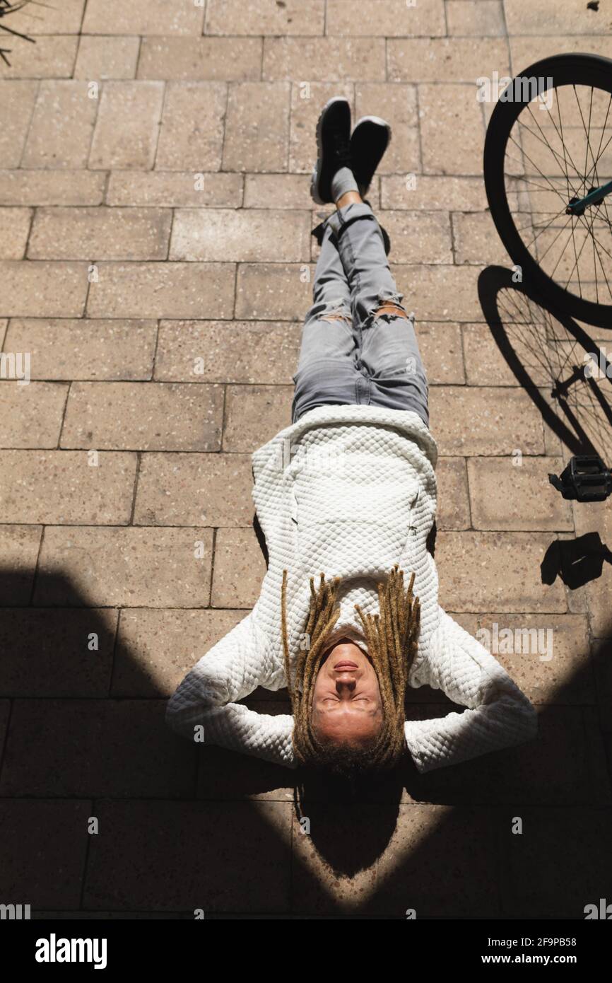 Mixed race male with dreadlocks lying on street with eyes closed and relaxing. green urban lifestyle, out and about in the city. Stock Photo