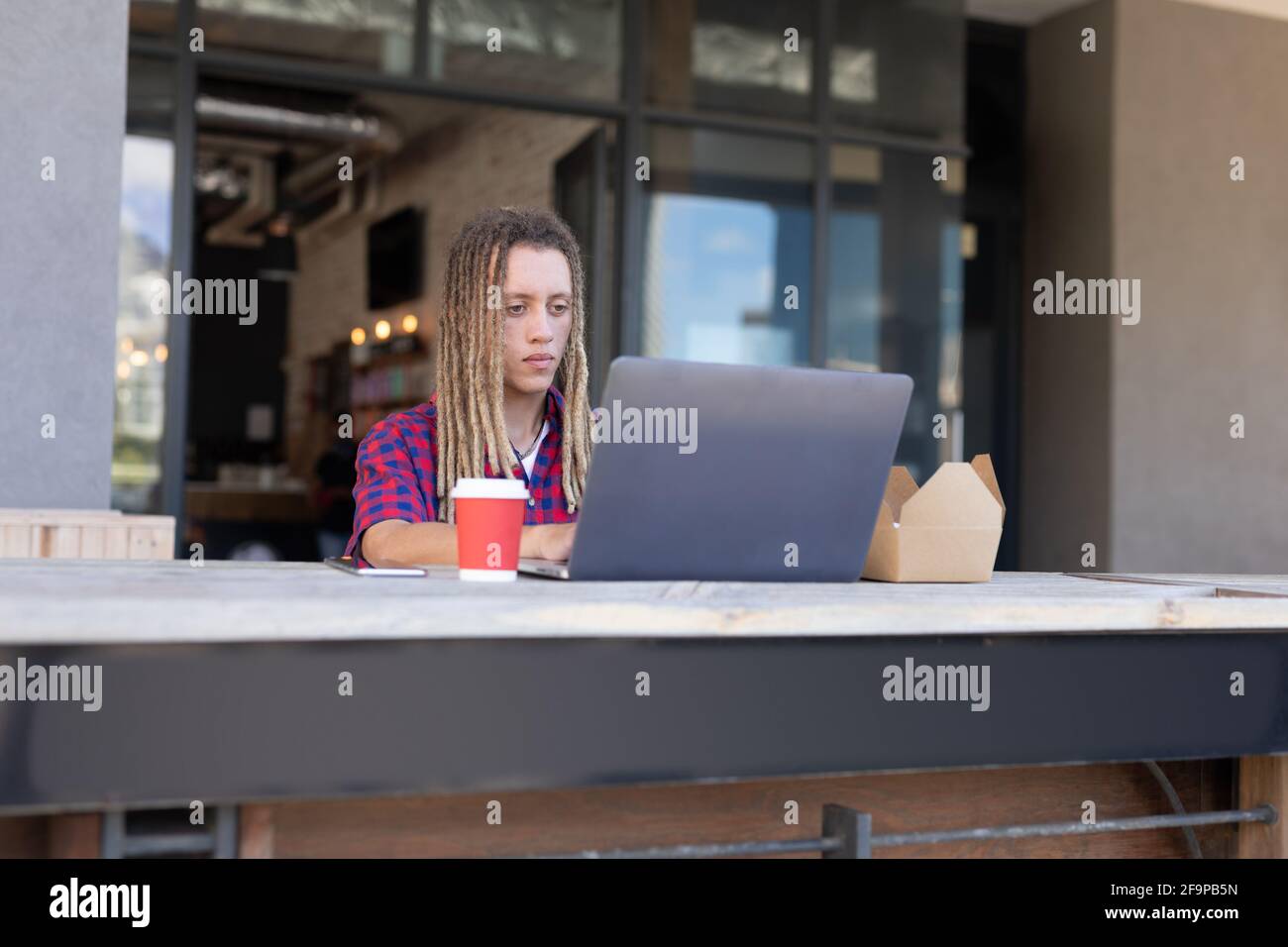 Mixed race man with dreadlocks sitting at table outside cafe using laptop. digital nomad, out and about in the city. Stock Photo
