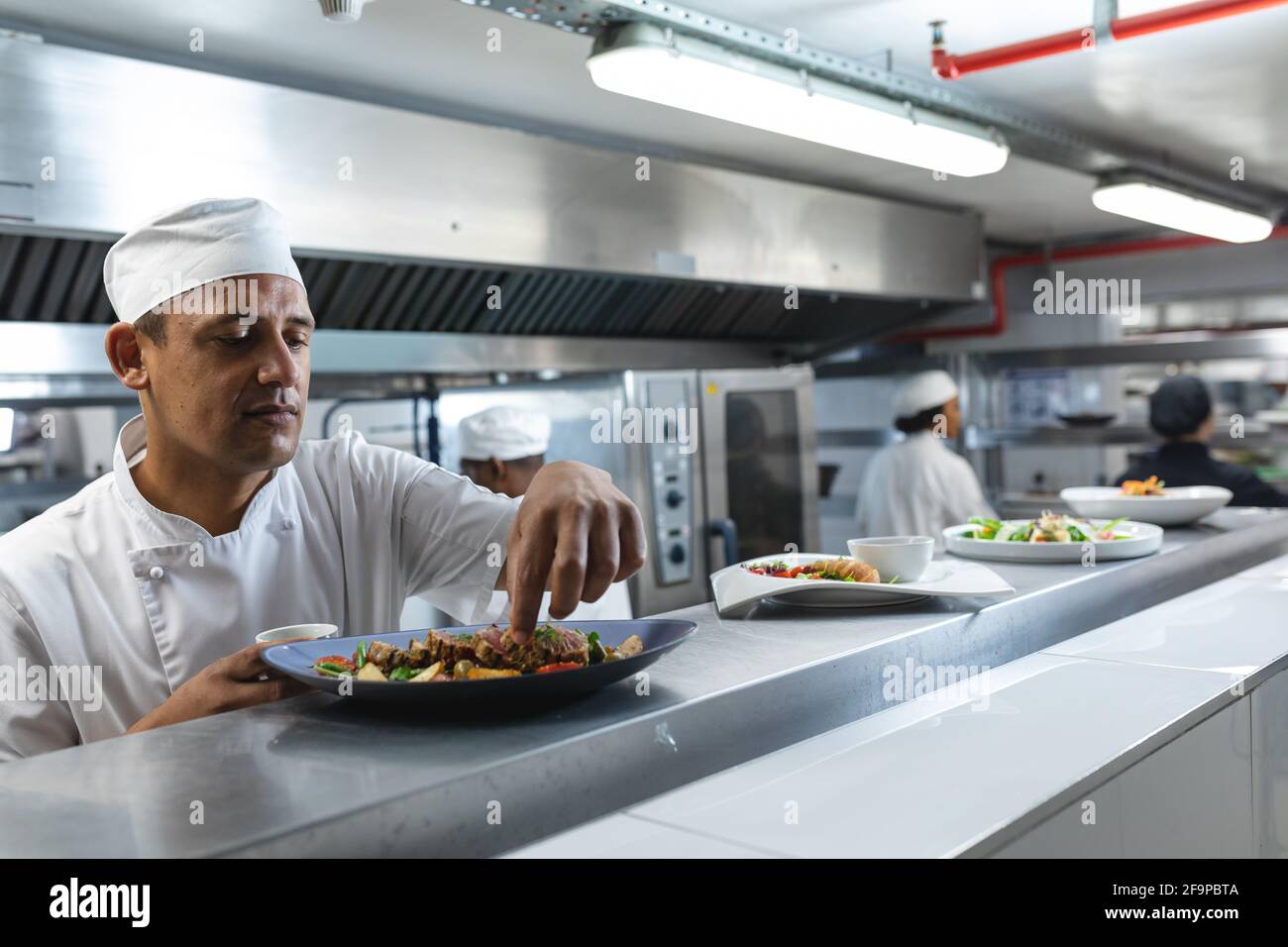 Mixed race professional chef finishing dish before serving with colleague in background. working in a busy restaurant kitchen. Stock Photo
