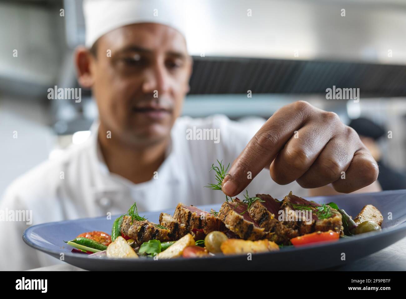Mixed race professional chef finishing dish before serving with colleague in background. working in a busy restaurant kitchen. Stock Photo