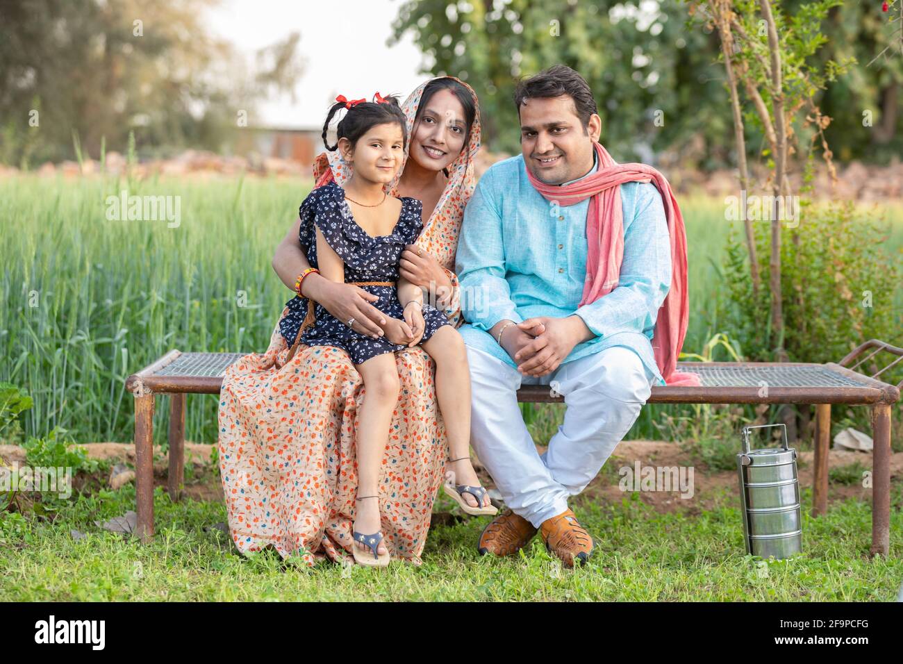 Happy Indian farmer family sitting on traditional bed at agricultural field in village, Young couple with their daughter in traditional outfit. Stock Photo