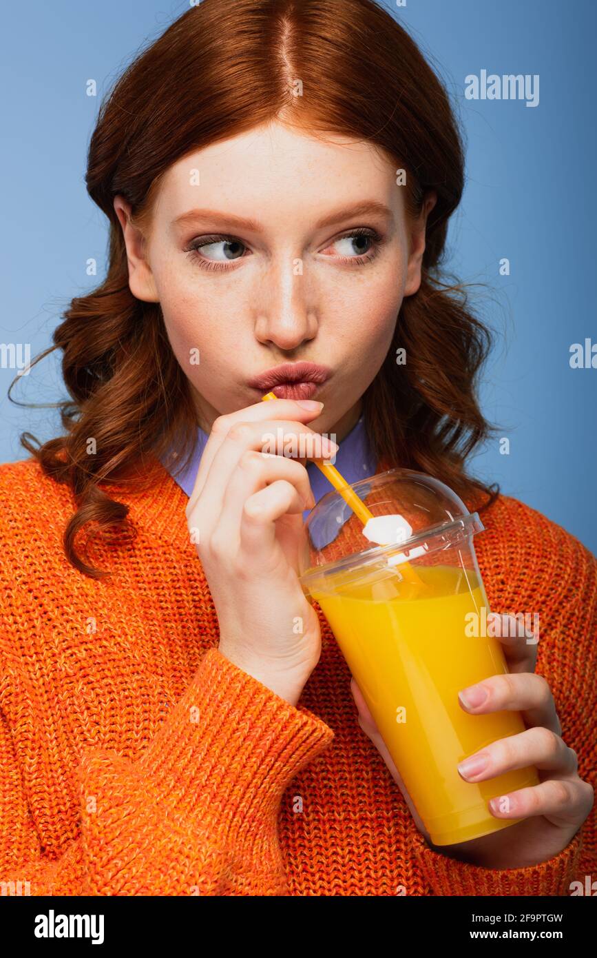 redhead woman in sweater pouting lips and holding fresh orange juice in