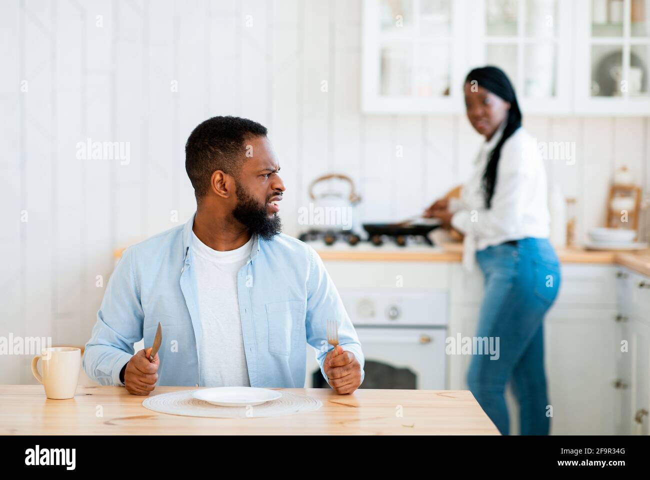 Impatient Hungry Black Husband Waiting For Food, Sitting At Table In Kitchen Stock Photo