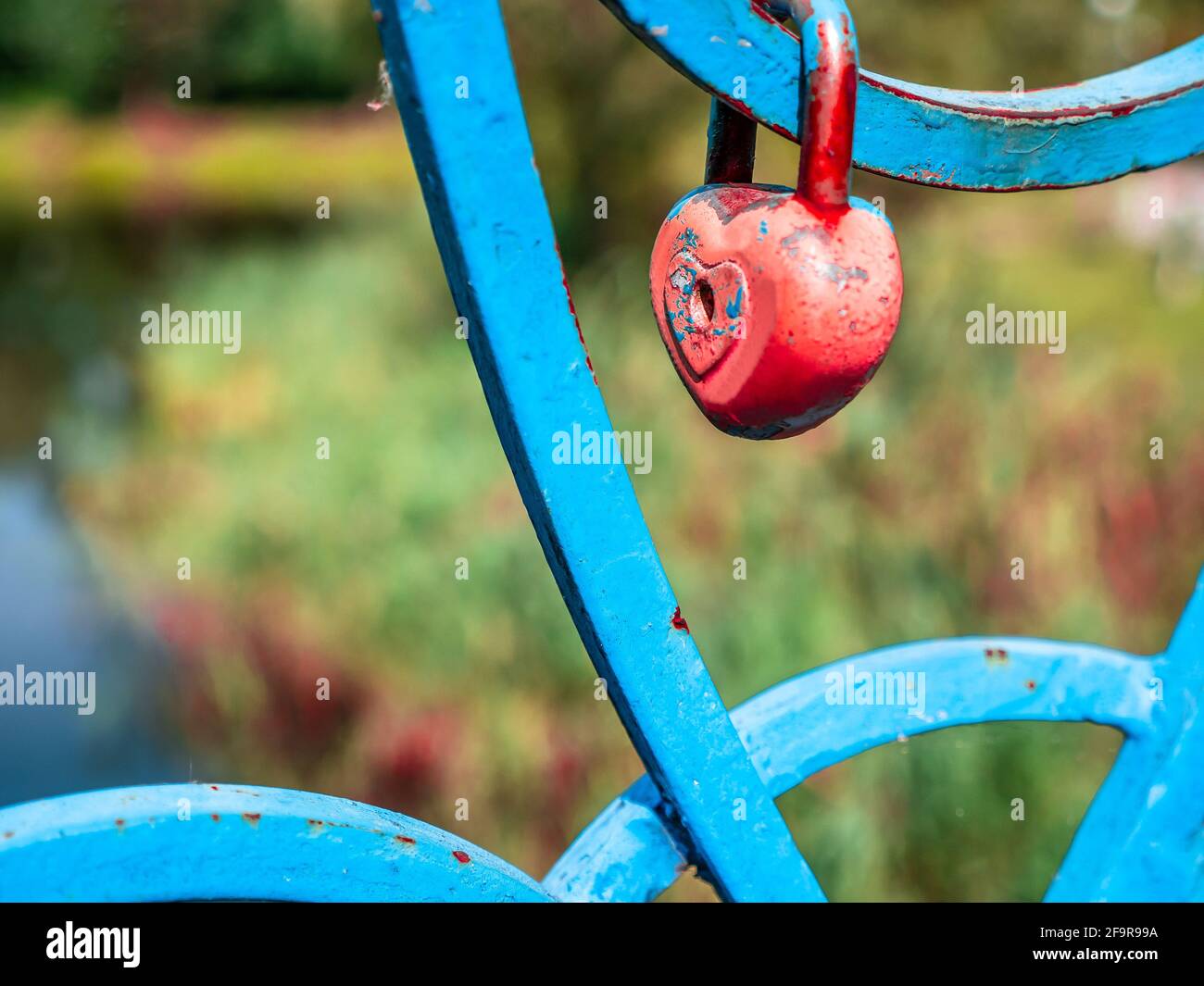 An old wedding lock in heart shape with soft edges and red peeling paint on a blue painted bridge fence under bright sunshine. Stock Photo