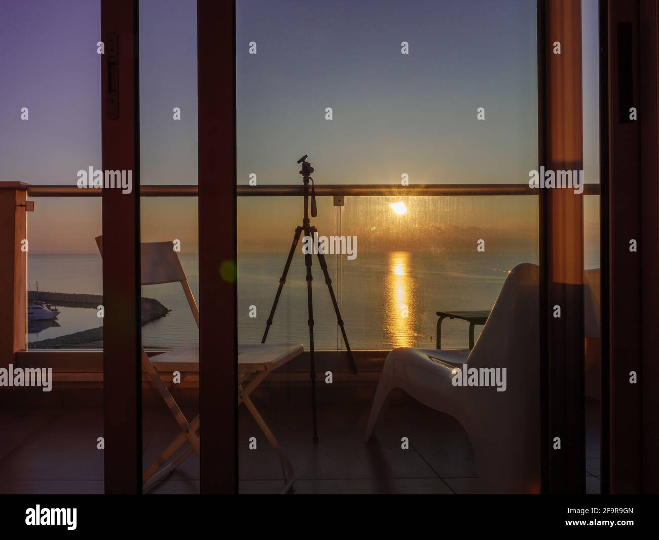 Tripod, chair, and chaise longue at a terrace with glass barrier and view over colorful sunset above the calm Mediterranean sea in Larnaca, Cyprus. Stock Photo