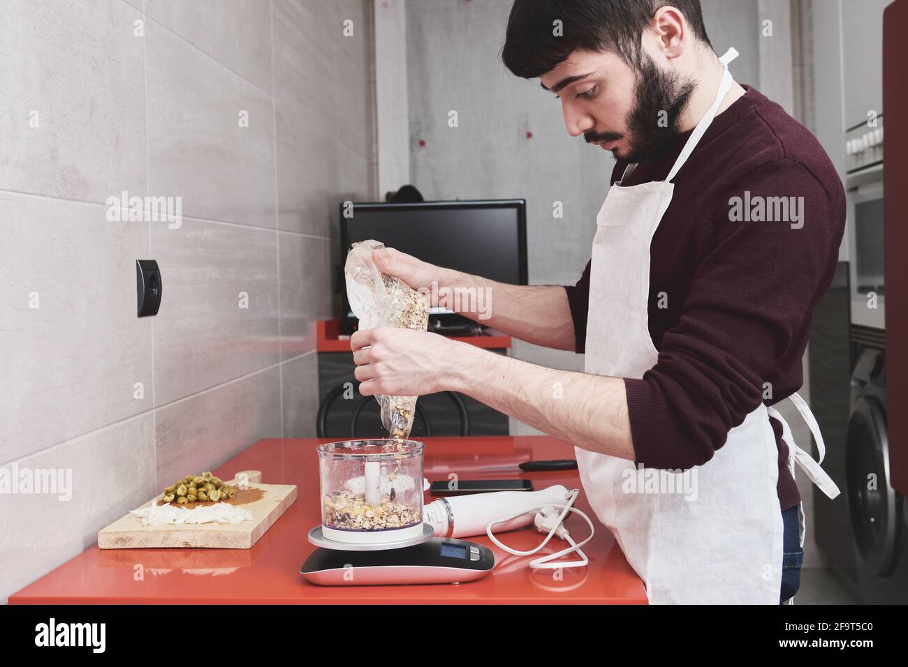 Young caucasian man in apron cooking in a kitchen. He is weighting muesli on a scale. Stock Photo