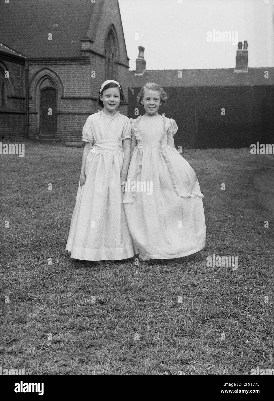 1956, historical, outside in the grounds of a church,standing for a photo,  two young girls in their pretty gowns they will wear in the traditional May Queen procession, England, UK. May Day is an ancient festival of Spring and celebrations , including Morris Dancing, the crowning of a May Queen and dancing around a Maypole having taken place in England for centuries. Selected from the girls of the area to lead the May Day parade, the May Queen wearing a crown, would start the celebrations and in the North of England, the Church Sunday Schools led the organisation of the day. Stock Photo
