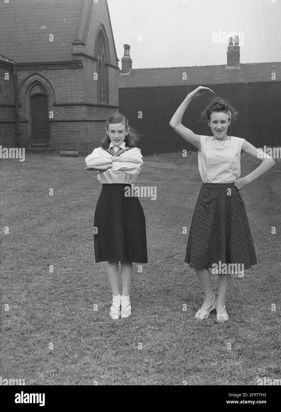 1956, historical, outside in the grounds of a church, two  girls in their outfits, showing the moves they will perform in the traditional May Queen carnival, England, UK. An ancient festival of Spring and celebrations, in many villages, May Day involved the crowning of a May Queen and dancing around a Maypole, activities that have taken place in England for centuries. Selected from the girls of the area, The May Queen wearing a crown, would start the procession of floats and dancing. In the industrialised North of England, the Church Sunday Schools often led the organisation of the day. Stock Photo