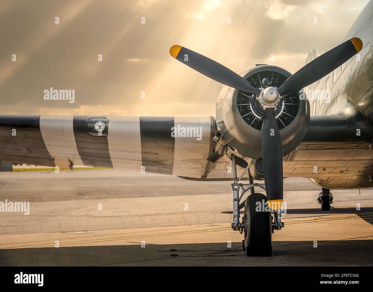 Old WW2 Dakota twin engine Operation Overlord aeroplane parked with sun setting over plane wings sunlight rays from sky. Wheel chocks three propellers Stock Photo