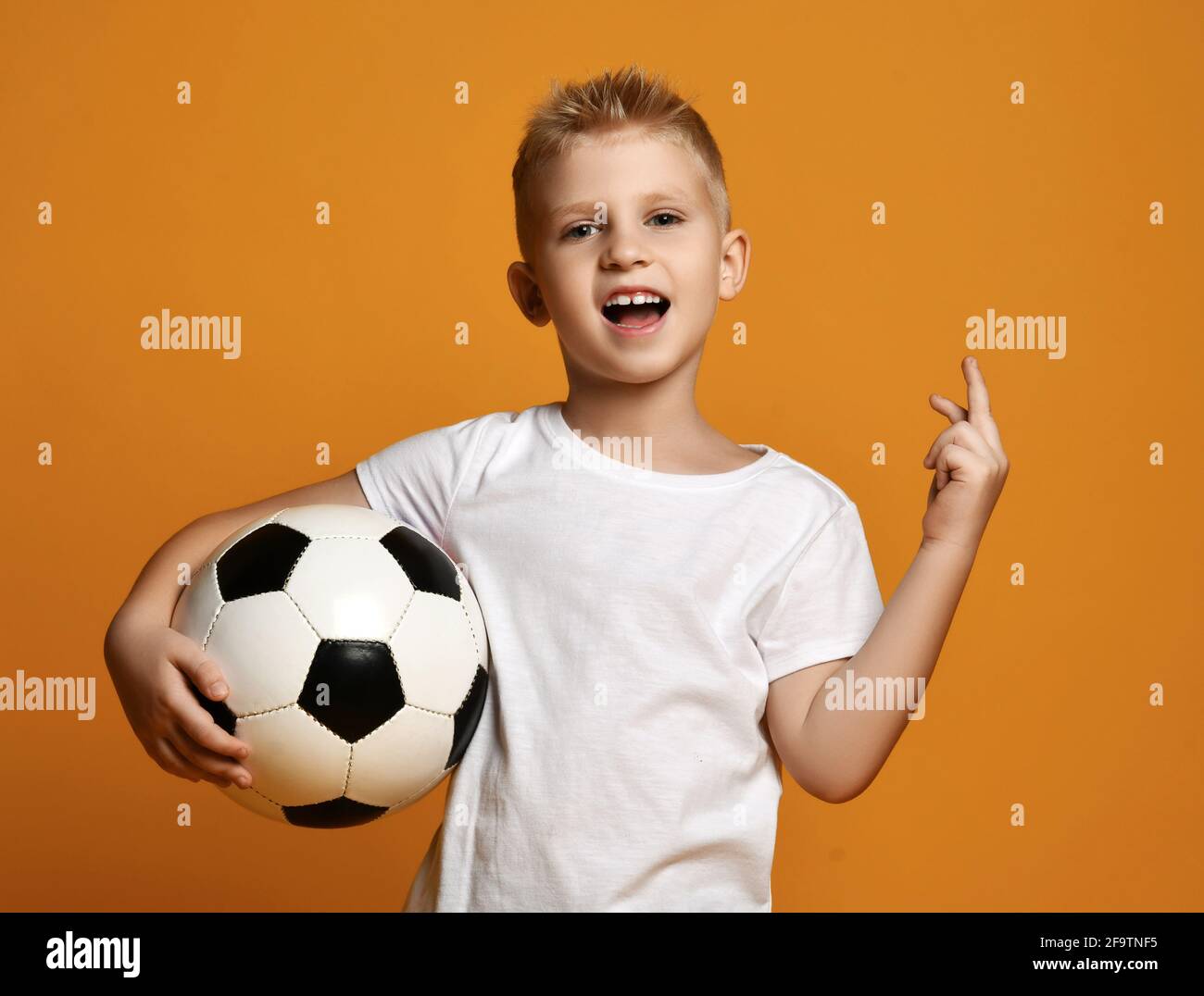 Active sporty blonde kid boy in white blank t-shirt stands holding soccer ball in hand and gestures one, first sign Stock Photo