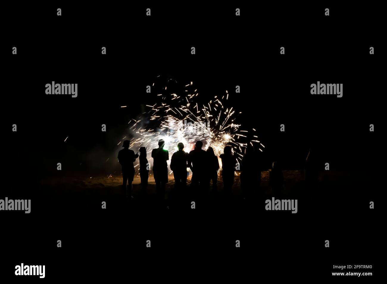 Group of people in silhouette watch fireworks explode on beach in total darkness. Stock Photo
