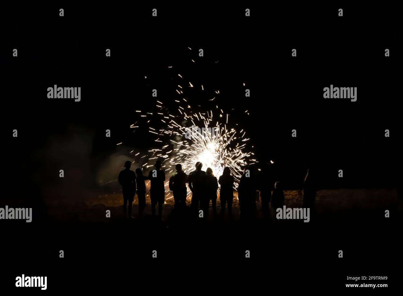 Group of people with backs turned in silhouette watch fireworks explode on beach at night. Stock Photo
