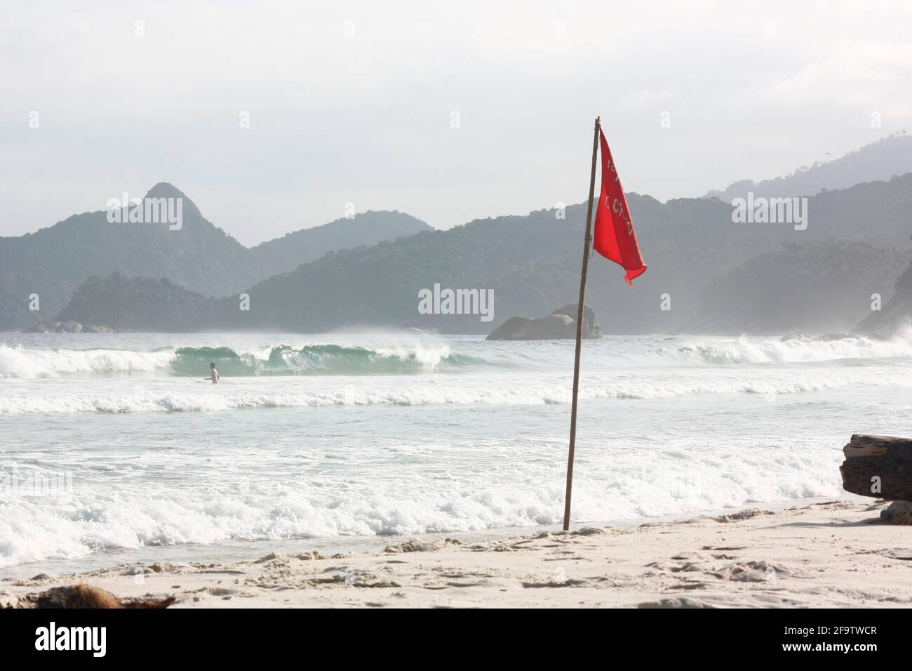 Lopes Mendes beach scene, Brazilian nature reserve. A red flag on the beach, a trunk, a sea, a fog and mountains. Stock Photo
