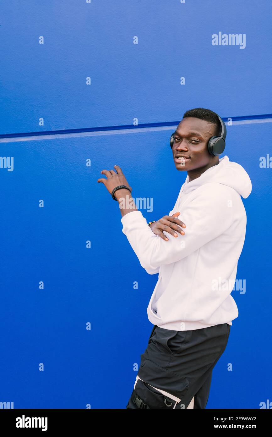 Young afro American black man wearing a white sweatshirt and headphones walking near a blue wall looking at camera. Modern look Stock Photo