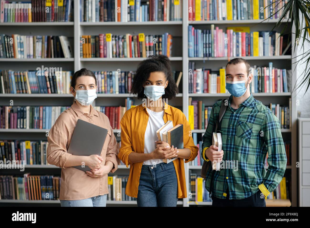 Education, student life, friendship. Multinational students friends, standing in the university library, wearing protective medical masks due to the coronavirus pandemic, looking at the camera Stock Photo