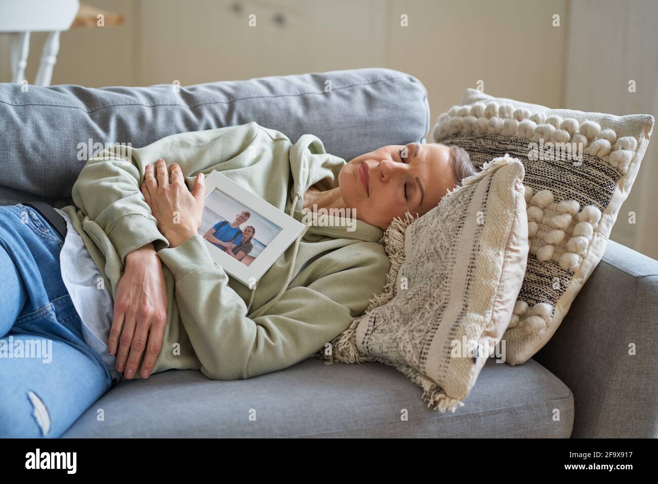 Sad middle aged woman with family photo with her children in frame keeping eyes closed and dreaming while lying on sofa at home. Selective focus on senior female Stock Photo