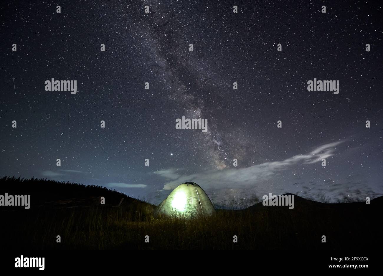 Amazing starry night sky in the mountains and illuminated tent at campsite. Millions of stars over single tent in the middle of mountain meadow. Concept of mountain retreat Stock Photo