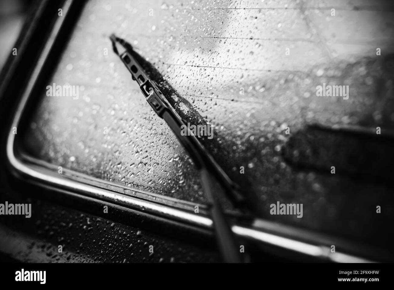 A black-and-white image of a rain-soaked car rear window with a dark windshield wiper. Cloudy, dreary weather. Transport. Stock Photo