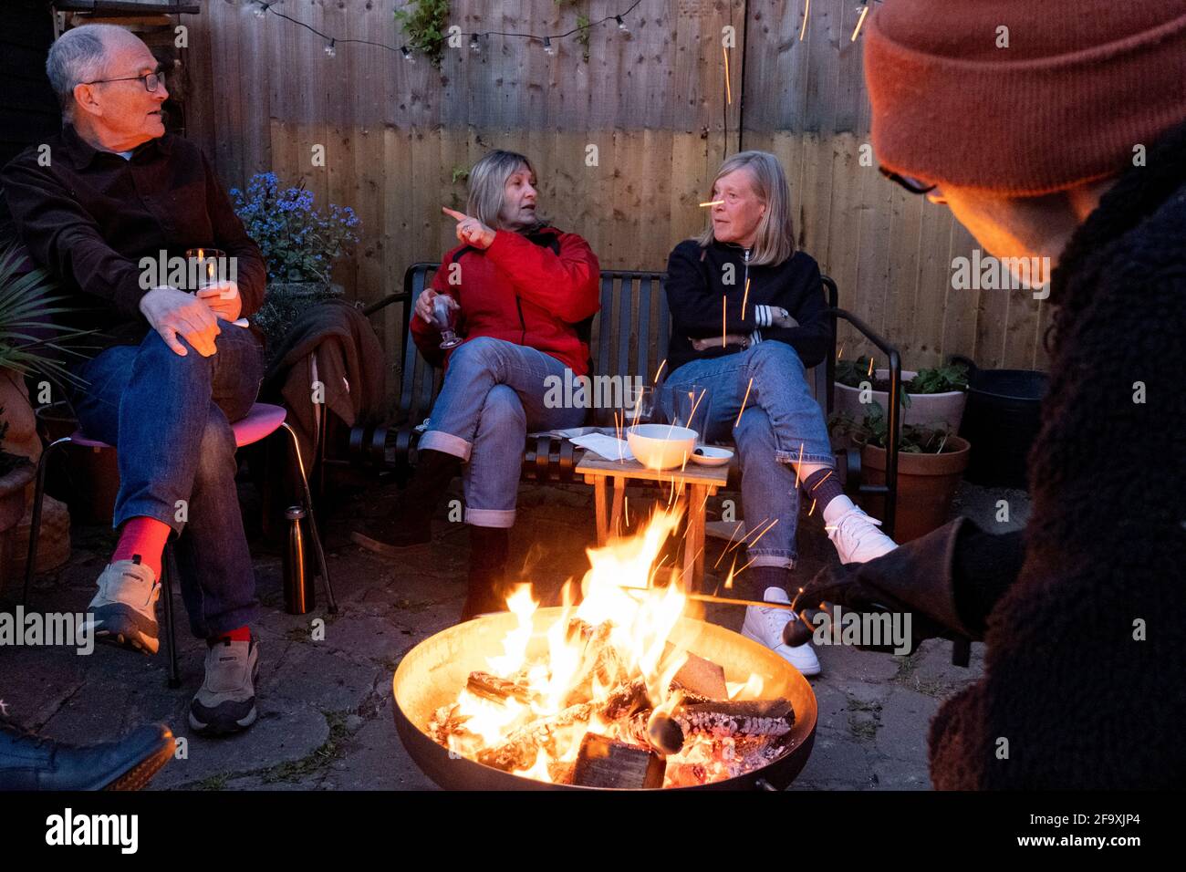 Friends catch-up with latest news and gossip while gathered around the warmt flames of a circluar fire bowl that hosts have lit in their south London back garden, on 17th April 2021, in London, England. Stock Photo