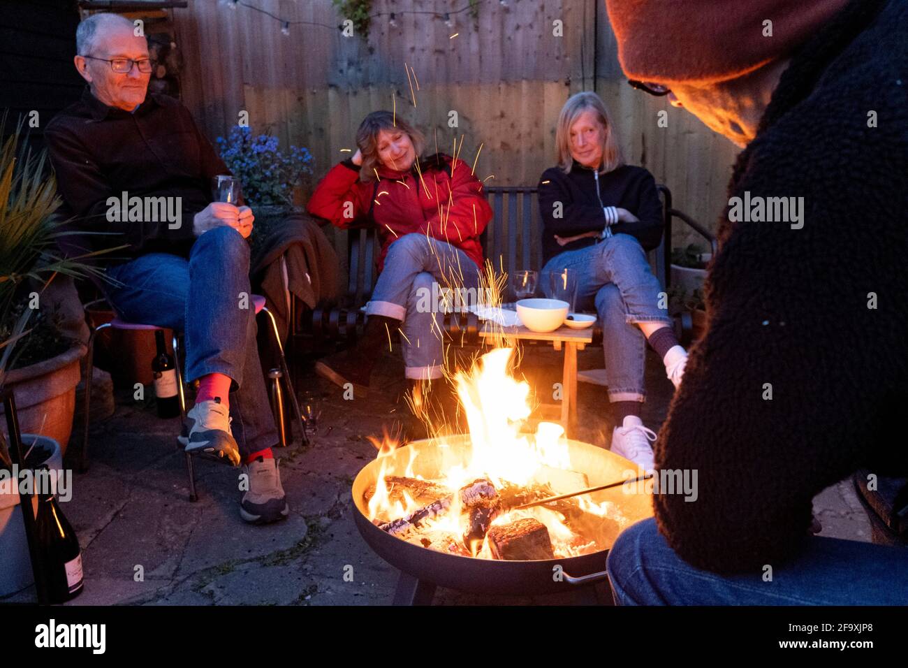 Friends catch-up with latest news and gossip while gathered around the warmt flames of a circluar fire bowl that hosts have lit in their south London back garden, on 17th April 2021, in London, England. Stock Photo