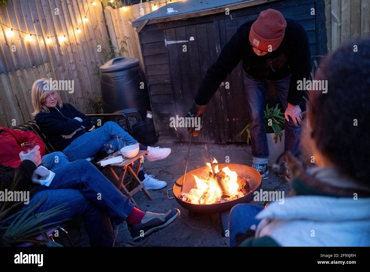 Friends catch-up with latest news and gossip while gathered around the warmt flames of a circluar fire bowl that hosts have lit in their south London back garden, on 17th April 2021, in London, England. Stock Photo