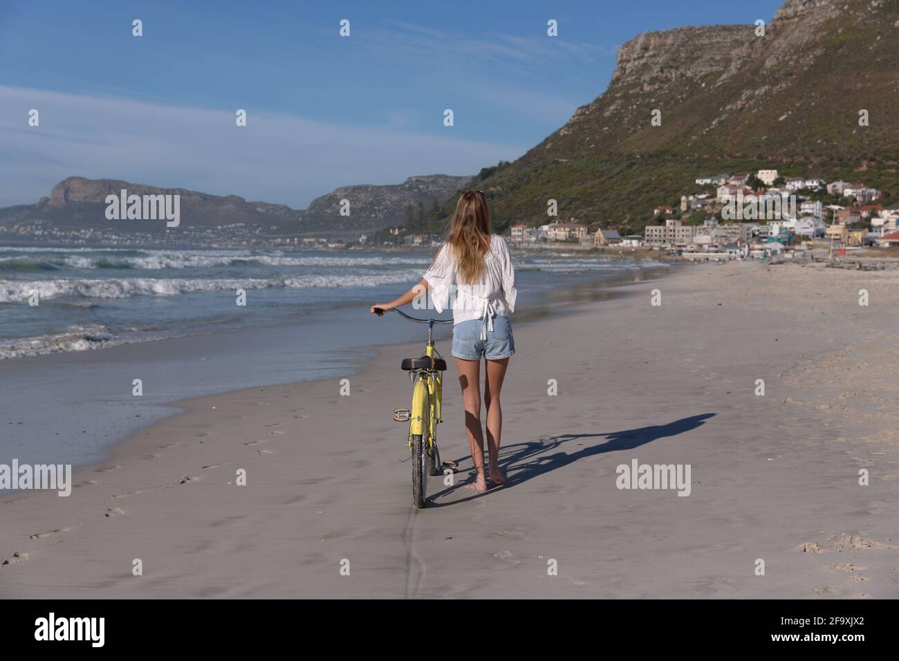 Rear view of caucasian woman in white top and shorts walking with a bicycle at the beach Stock Photo