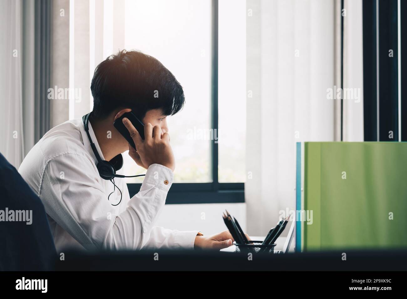 Side view of young man entrepreneur in office making phone call while working with laptop. Stock Photo