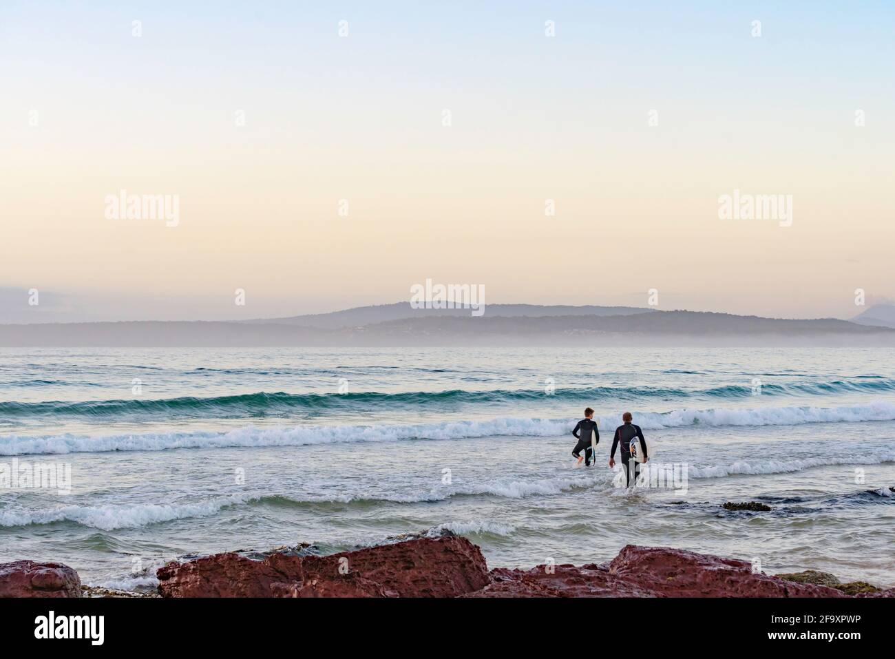 A father and son wade out in dawn mist with their boards towards the surf at Bar Beach in Merimbula on the New South Wales south coast of Australia Stock Photo