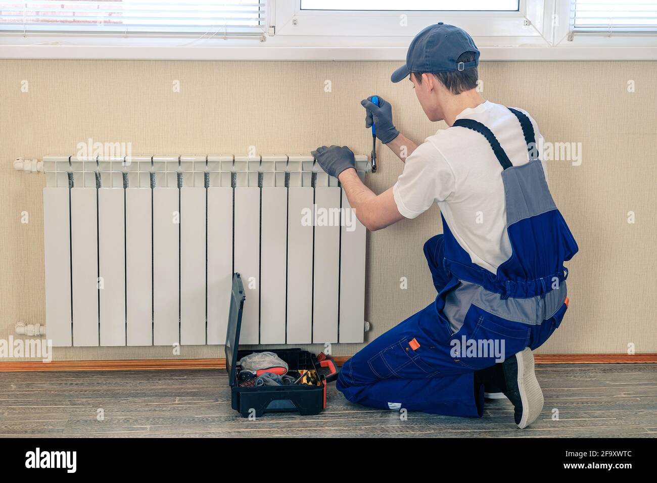 A plumber in a blue jumpsuit is fixing a radiator. With an adjustable wrench, he tightens the tap. A briefcase with a tool at hand Stock Photo