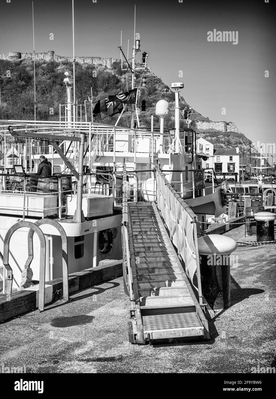 A boat moored in a harbour with a gangway leading up.  A person sits on the upper deck and an ancient castle wall is in the background. Stock Photo