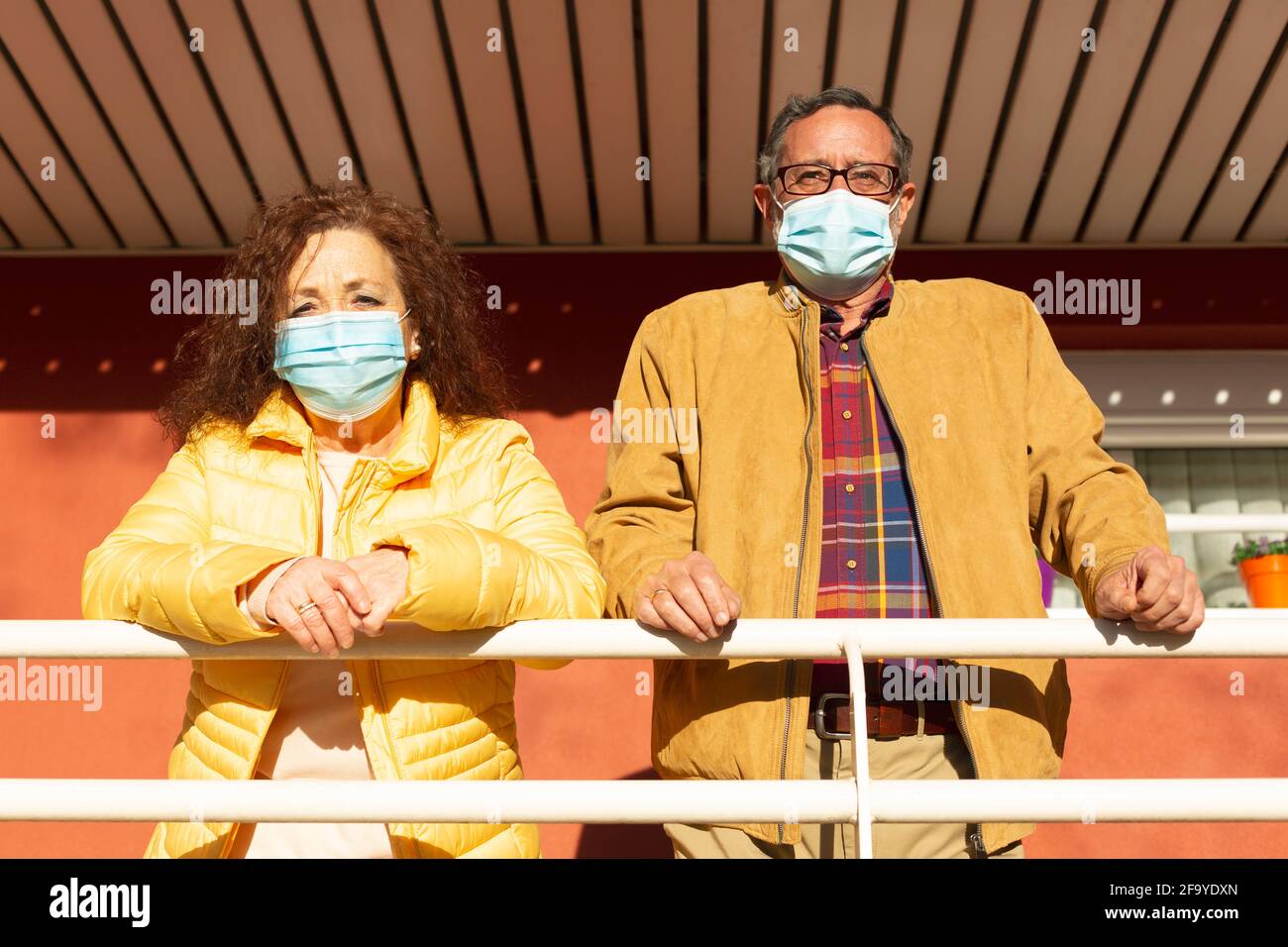 Portrait of two senior citizens observing the outside from the porch of their home. They are wearing a protective mask to prevent the Covid-19 virus. Stock Photo