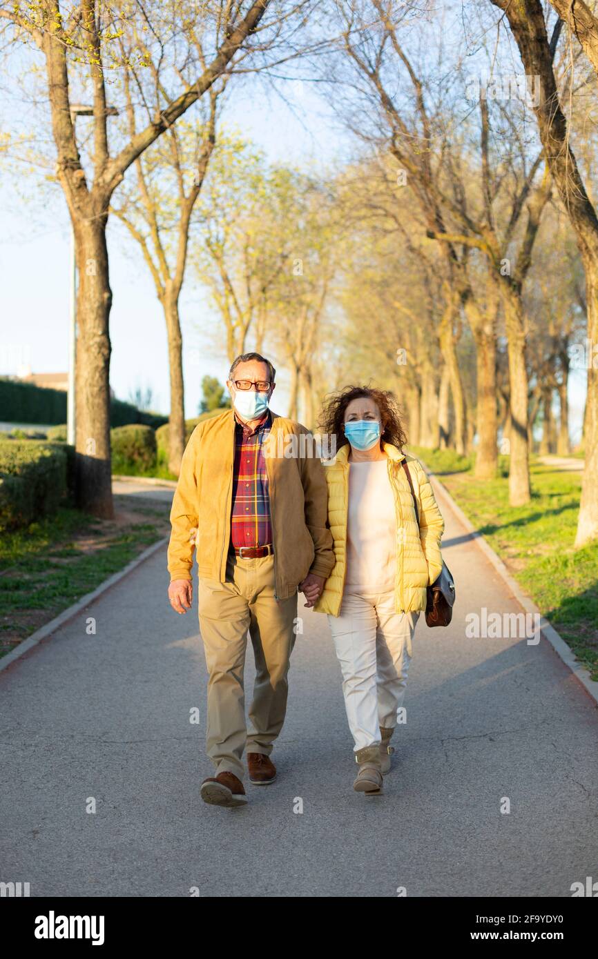 Senior couple walking outdoors holding hands. They are wearing a surgical mask as a protective measure against the Coronavirus. Space for text. Stock Photo