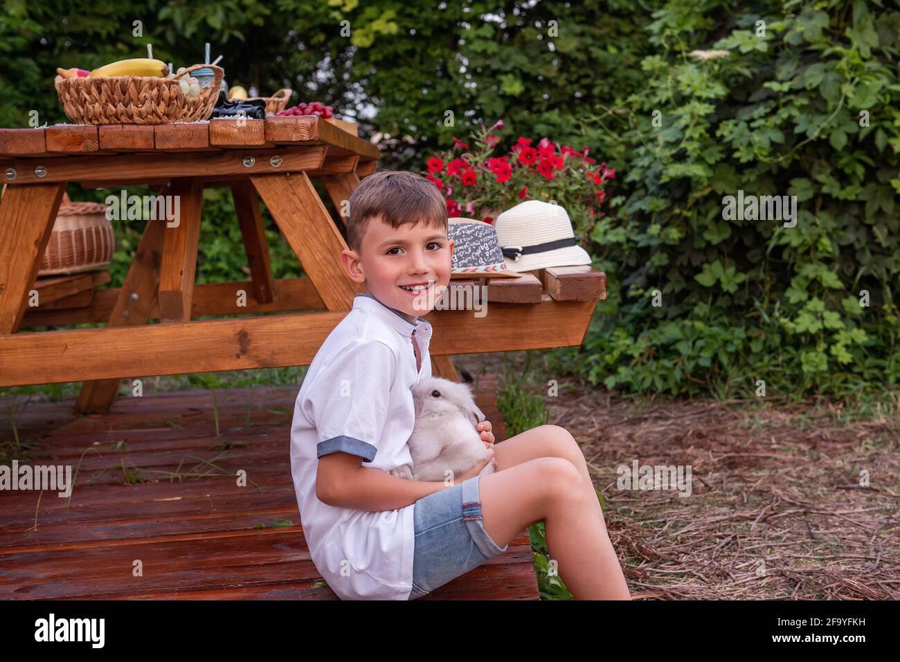Portrait of a little boy in a straw hat sits near a trailer truck at home on a wooden floor, holding a fluffy rabbit in his hands. Laughs, plays with Stock Photo