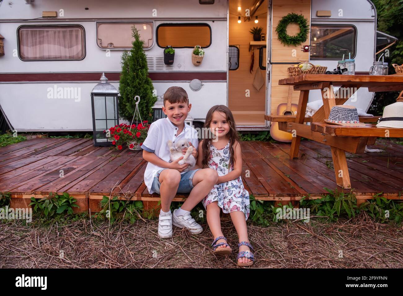 Boy and girl are sitting at trailer truck home on wooden floor, holding fluffy rabbit bunny in their hands. Brother sister hug, play with domestic har Stock Photo