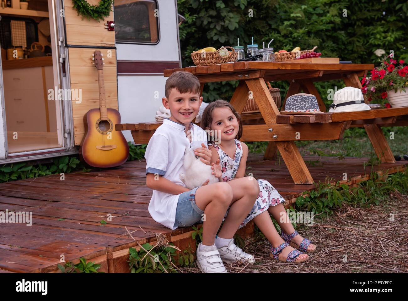 Boy and girl are sitting at trailer truck home on wooden floor, holding fluffy rabbit bunny in their hands. Brother sister hug, play with domestic har Stock Photo