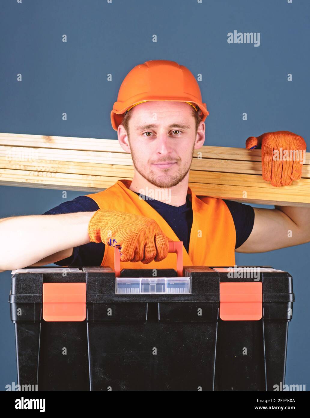 Man in helmet, hard hat holds toolbox and wooden beams, grey background. Professional woodworker
