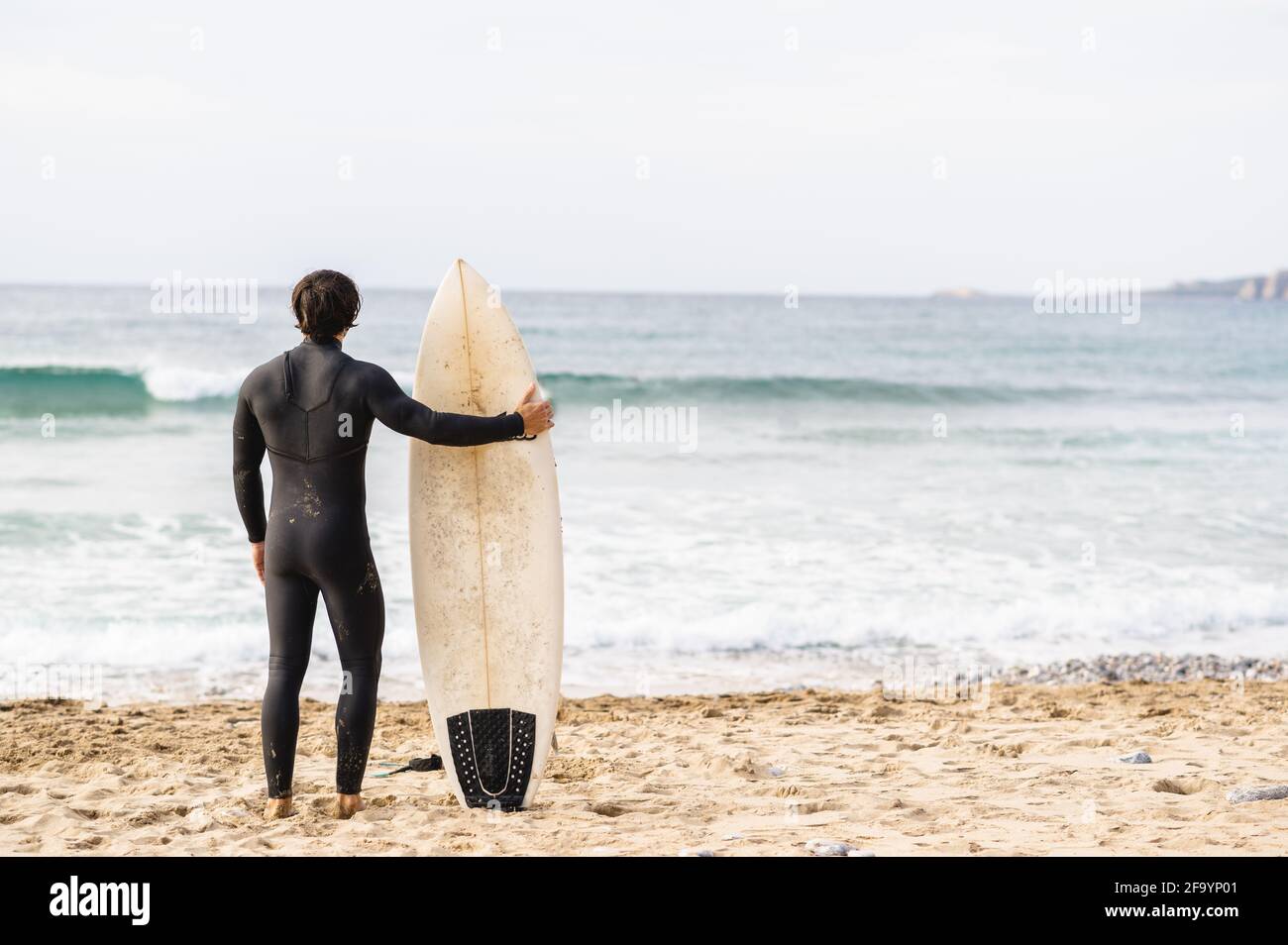 Back view of surfer man wearing diving suit leaning on the surfboard