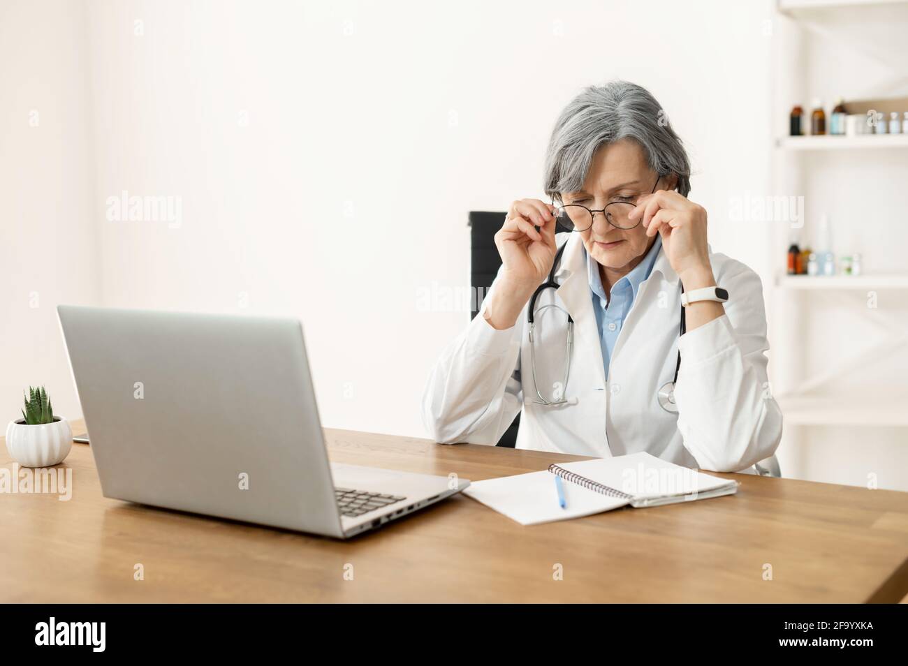 Senior old female doctor with stethoscope in a lab coat and glasses is done with work, removing her glasses, sitting at the desk and looking at the notebook, studying online, freshening-up her memory Stock Photo