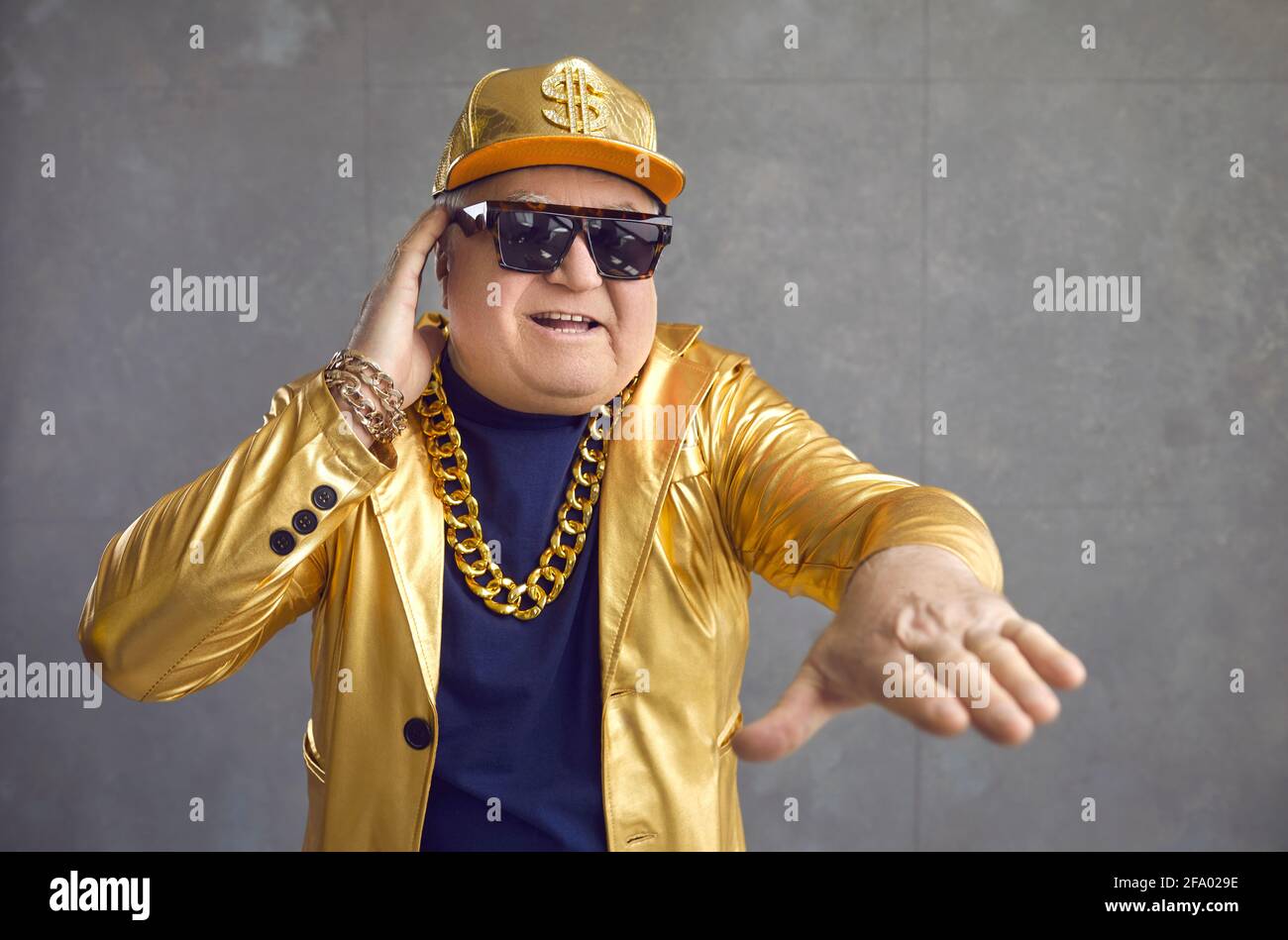 Funny senior man in golden jacket, baseball cap and chain necklace playing music at disco party Stock Photo