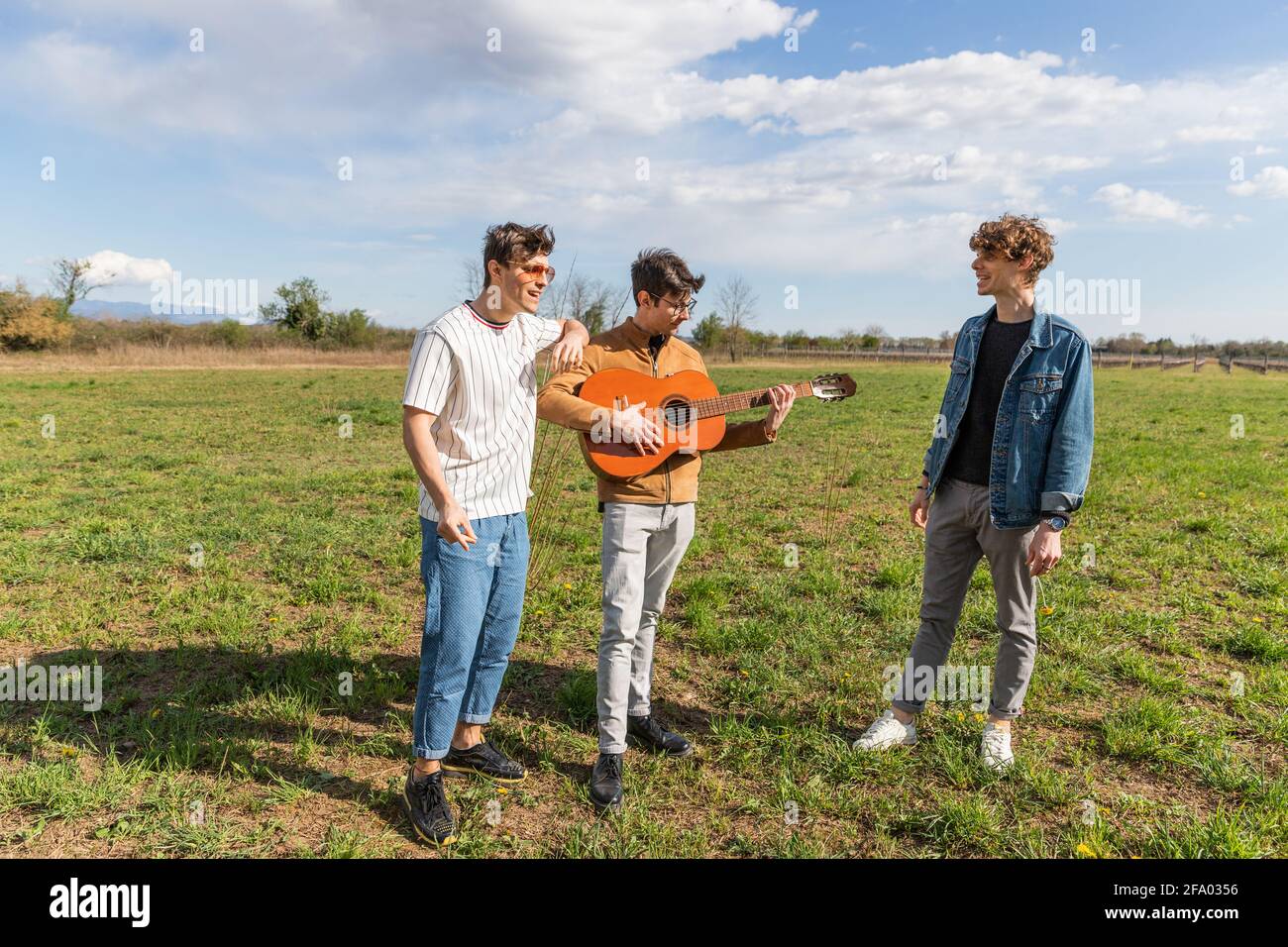 three friends in a field meet to sing and play guitar cheerfully together. Stock Photo