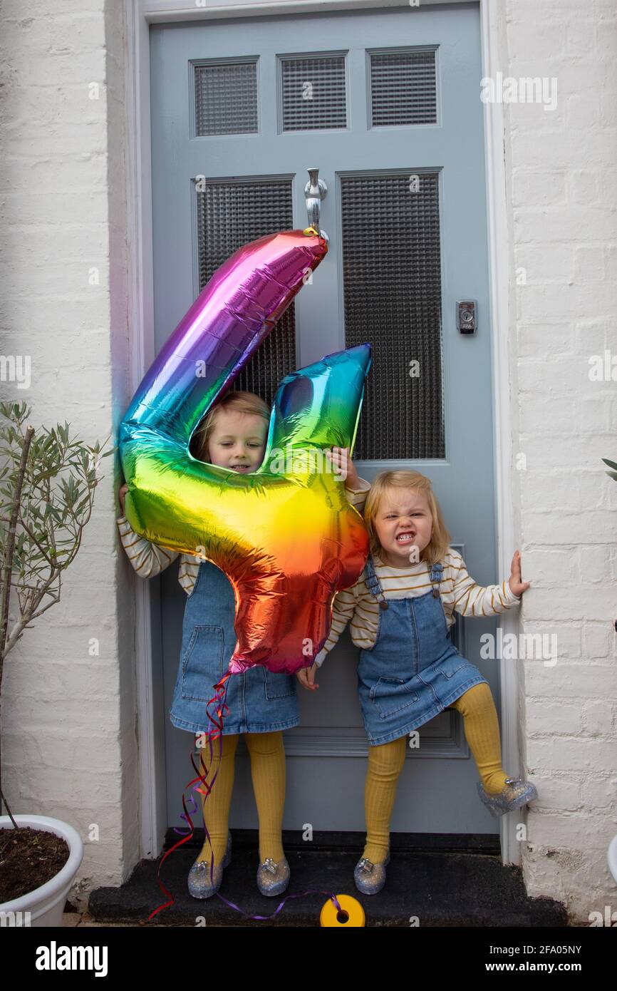 Young sisters holding a large rainbow coloured helium no 4 balloon outside their front door in the UK during lockdown. Stock Photo