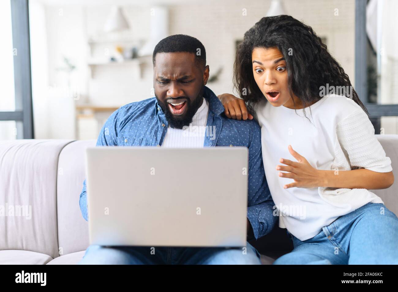 Vexed and surprised African-American couple sits on the couch at home and looks at the laptop screen, received unexpected news, lost in video game Stock Photo