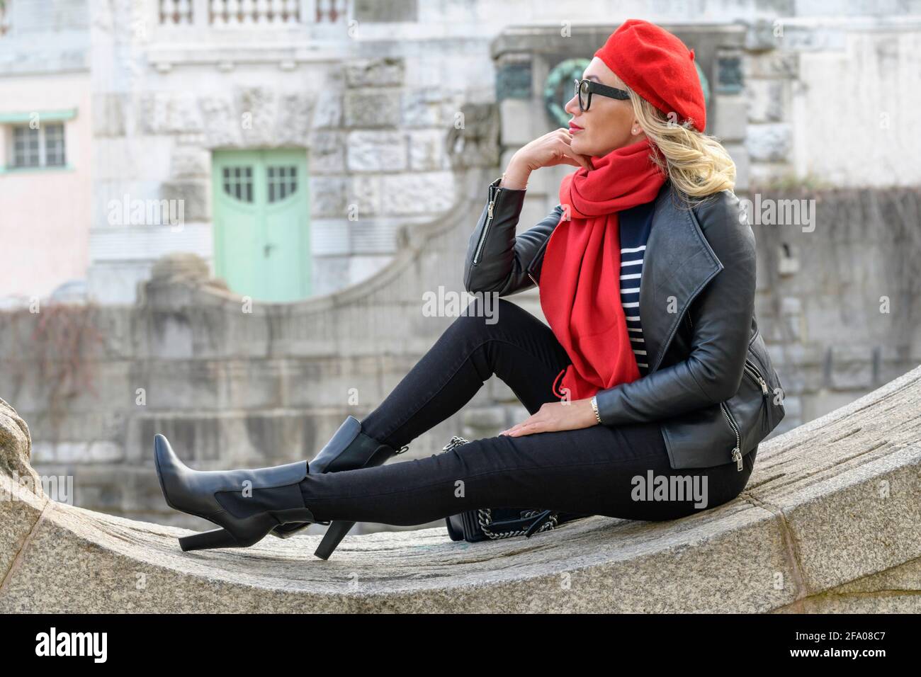 Blonde lady with a red scarf and hat and a black leather jacket is sitting  on a wall Stock Photo - Alamy