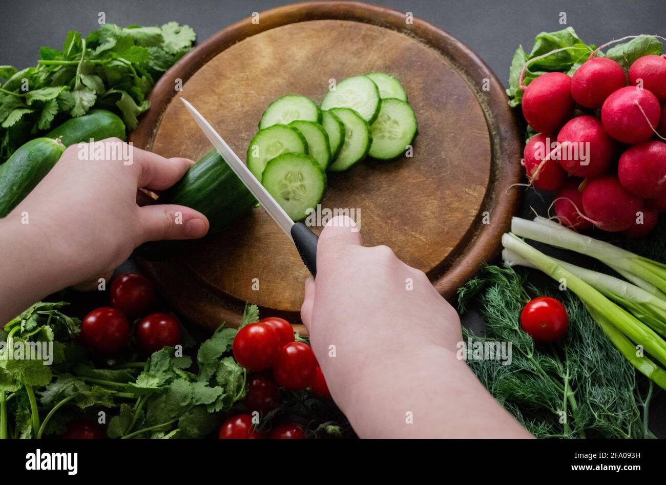 Hands cutting cucumber with a white ceramic knife on a round wooden chopping board. Around the board are ripe fresh vegetables: tomatoes, onions, radi Stock Photo