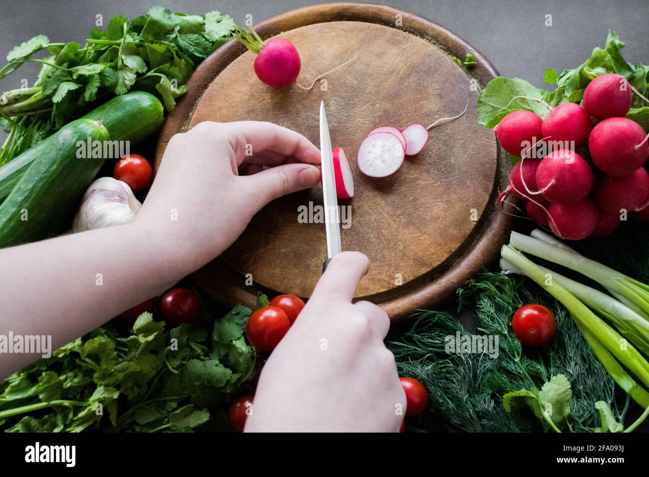 Hands cutting radishes with a white ceramic knife on a round wooden chopping board. Around the board are ripe fresh vegetables: tomatoes, onions, cucu Stock Photo