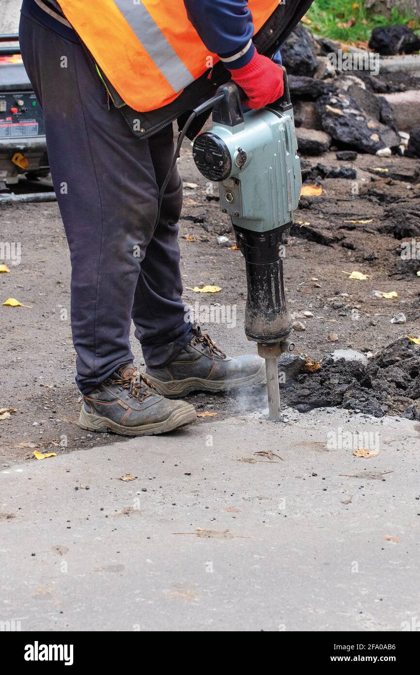 A road worker wearing a reflective jacket smashes old asphalt off the road with an electric jackhammer. Vertical image, close-up. Stock Photo