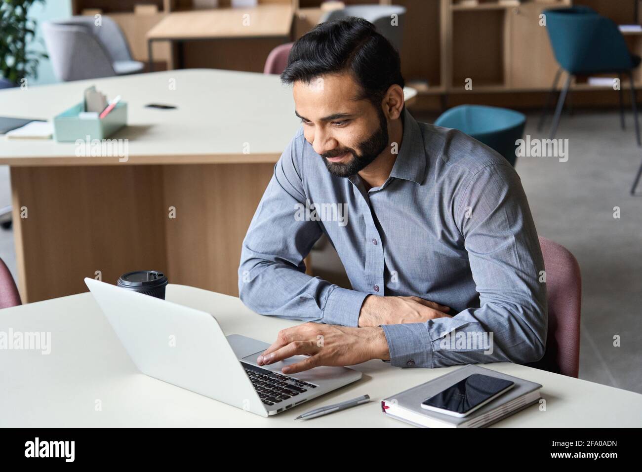 Smiling indian business man working with laptop and cellphone in coworking. Stock Photo